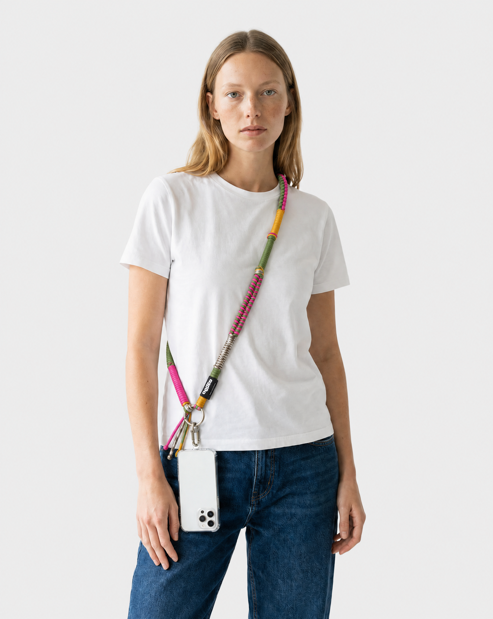 A woman wearing a white t-shirt and blue jeans models a colorful beaded crossbody phone strap with a phone attached, set against a plain light gray background.